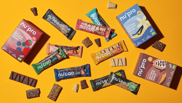 A colorful assortment of health food products is displayed on an orange background. There are nu+pro and nu+seed boxes, along with various flavors of nu*cao bars. Some bars are unwrapped, showing their texture. Small bite-sized pieces are scattered around.