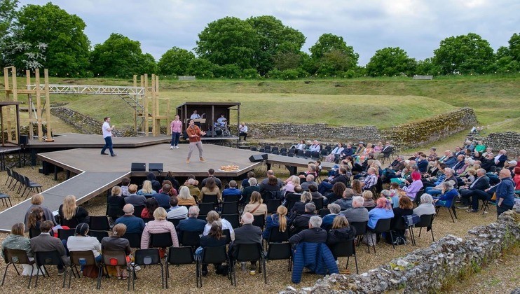 An outdoor theater performance is taking place in front of an audience seated on chairs. The stage is surrounded by grassy areas and old stone walls. Several actors are performing energetically under a cloudy sky with lush green trees in the background.