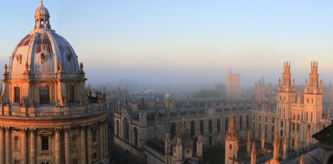 A panoramic view of Oxford University's Radcliffe Camera and surrounding historic buildings bathed in soft morning light. The sky is clear, and the distant horizon is a bit foggy, adding a mystical quality to the scene. The architecture is grand and elegant.