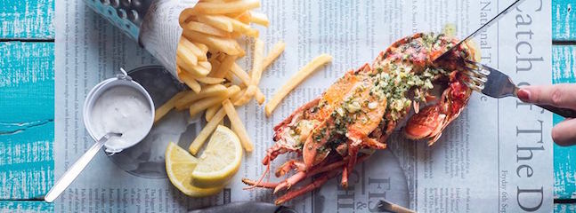 A meal featuring a lobster topped with herbs and garlic butter, served alongside a portion of golden french fries and a lemon wedge on a rustic blue wooden table. A side of creamy dipping sauce and a metal container holding extra fries are also visible.