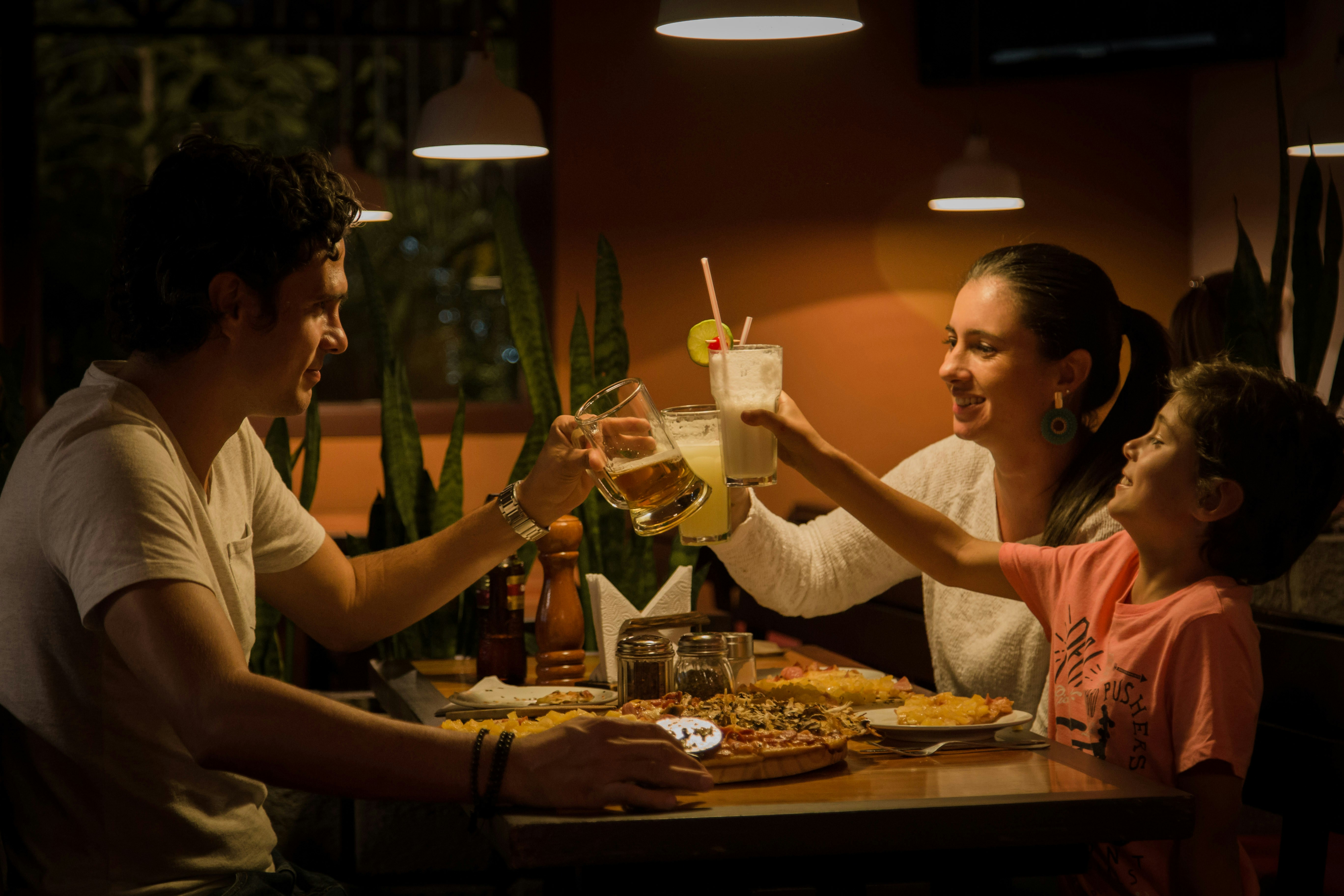 Male, female and child sitting and holding up drinks, in dimly lit restaurant at a table