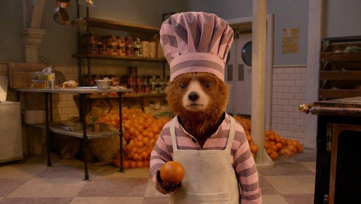 A bear dressed as a chef, wearing a striped pink and white chef hat and apron, stands in a kitchen holding an orange. The kitchen has shelves filled with jars and sacks of oranges piled on the floor.