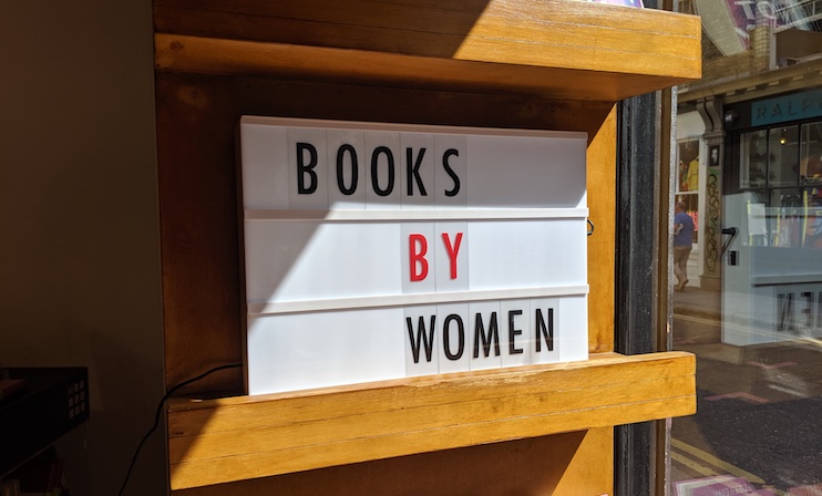 A lightbox sign on a wooden shelf displaying the message BOOKS BY WOMEN with the word BY highlighted in red. The sign is situated near a window, allowing natural light to highlight the display.
