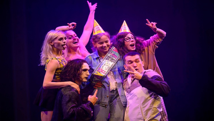 A group of six people posing for a photo on stage. They are dressed in various costumes, with some wearing party hats. One person is holding a colorful electronic device. Several members are making peace signs and playful expressions. The background is dark.