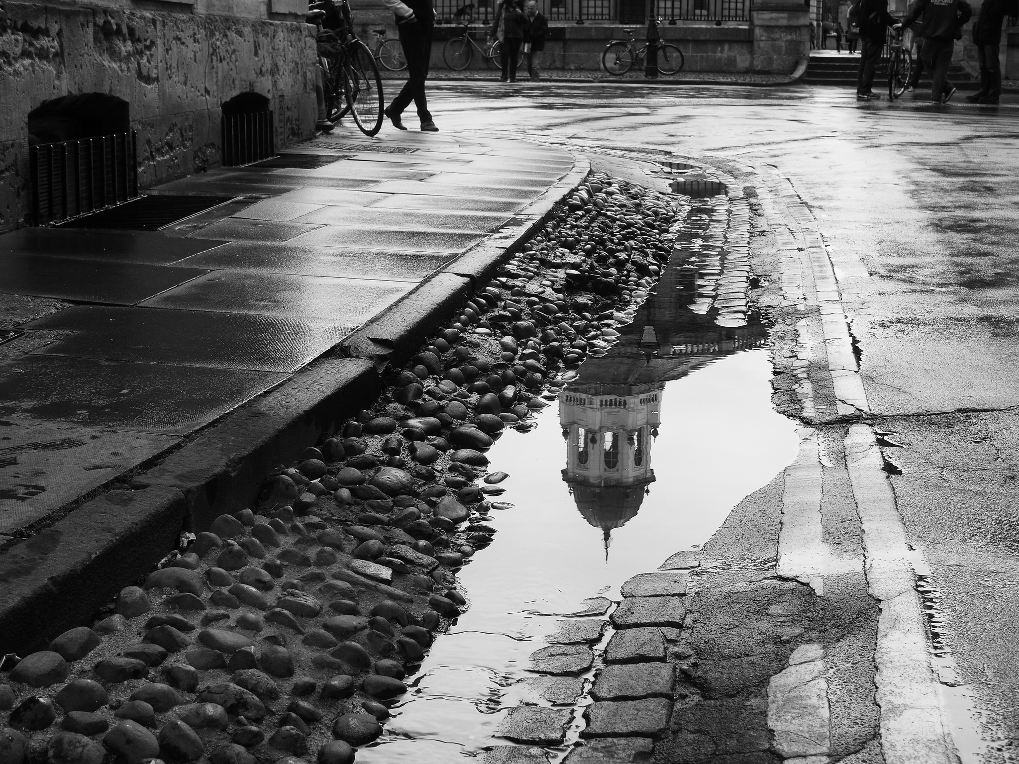 A black and white image of a wet cobblestone street with puddles. One puddle reflects the top of a historic building with a dome, capturing the architectural detail. People are seen walking and bicycles are parked along the side. The scene suggests recent rain.
