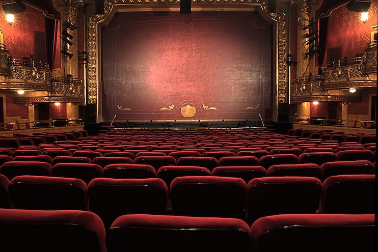 A lavish theater with ornate gold decorations and rows of red velvet seats facing a grand stage with a closed red curtain. Empty balconies flank the stage on both sides, and the entire space is illuminated with warm, classic lighting.