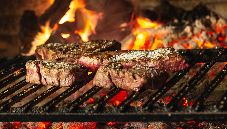Four pieces of seasoned meat are cooking on a grill over an open flame with glowing embers underneath. The meat appears to be searing with a slight char, giving it a savory appearance. The background features a fiery, rustic setting.