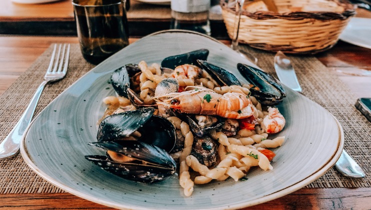 A plate of seafood pasta is served on a triangular dish, featuring mussels, shrimp, and twisted pasta noodles. The plate is set on a woven placemat with a fork, knife, and a glass nearby. A wicker basket is visible in the background.