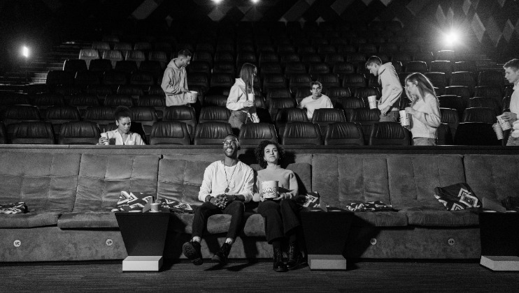 Black and white photo of a nearly empty movie theater. A couple sits in the front row, holding snacks. In the background, a few other people are scattered across the seats, some holding popcorn. The theater is dimly lit with a visible spotlight.