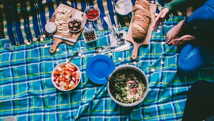 A top-view photo of a picnic setup on a blue plaid blanket. It features bowls of salad and fruit, a loaf of bread, crackers, cheese, assorted toppings, and several utensils. Two people are sitting with their legs crossed, partially visible on the right side.