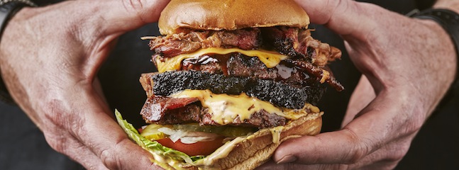 Close-up of a pair of hands holding a large, stacked barbecue sandwich. The sandwich consists of layers of shredded meat, thick-cut brisket slices, melted cheese, lettuce, and tomato on a toasted bun.