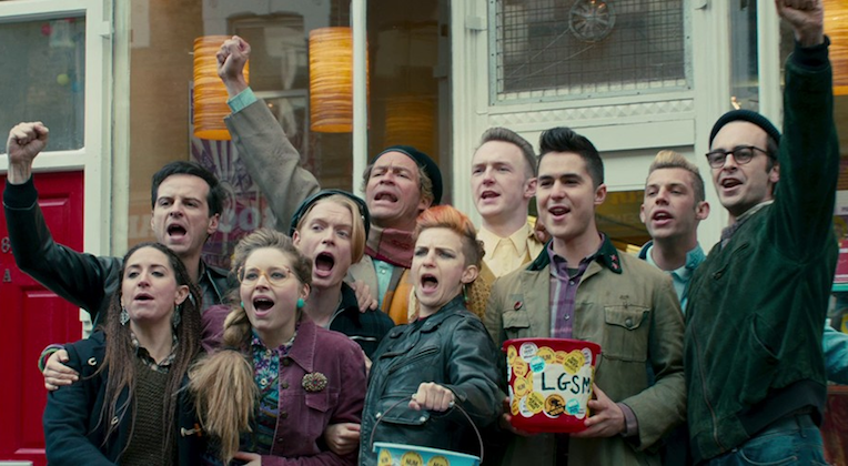 A group of nine people, composed of men and women, stand together outside a building with yellow lamp decorations inside. They appear to be celebrating, with some raising their fists and others holding containers labeled with stickers and LGSM.