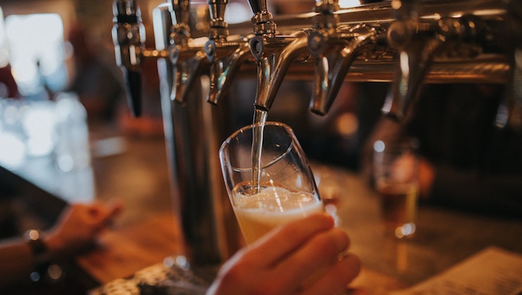 A person fills a glass with beer from a row of taps in a bar. The scene captures a dimly lit, warm atmosphere with another beer glass and patrons visible in the background. The beer is light in color and foamy at the top.