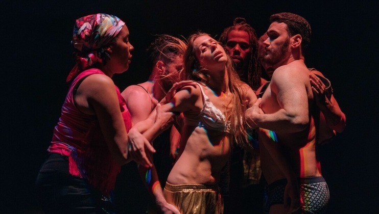 A group of dancers in various costumes are on stage performing. One person in the center, wearing a white top and gold pants, is being supported by the others. The lighting casts colorful shadows on their bodies. The background is dark.