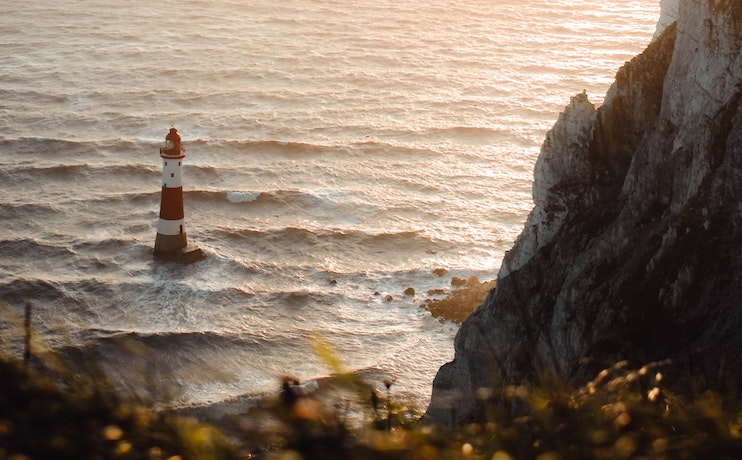 A red and white striped lighthouse stands in the sea near rugged cliffs at sunset. The waves crash against the base of the lighthouse, and the warm golden light of the setting sun reflects off the water and rocks, creating a serene and dramatic coastal scene.