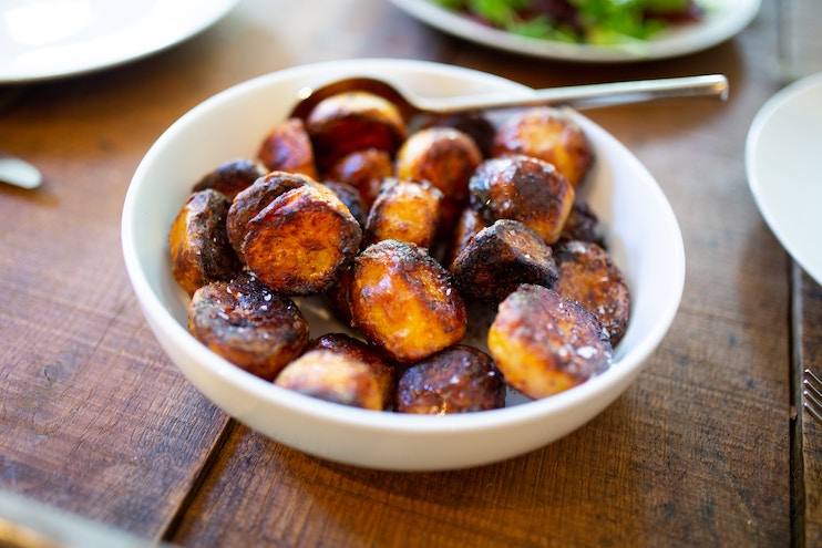 A white bowl filled with golden-brown, crispy roasted potatoes is placed on a wooden table. A spoon rests in the bowl. Other plates with food are blurred in the background. The potatoes have a slightly charred, caramelized exterior.