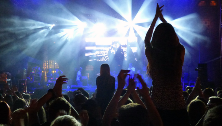 A lively concert scene with bright stage lights illuminating a crowded audience. People are clapping and cheering, and some are standing on their seats. The stage features performers and large screens displaying visuals in the background.