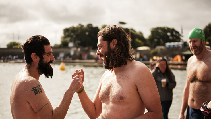 Two shirtless men with wet hair shake hands and smile at each other near a body of water. One man has a tattoo on his upper arm and the other has longer hair. In the background, people and trees are visible, suggesting a lively outdoor event or swim gathering.