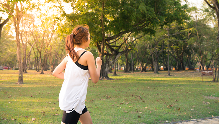 A person with a ponytail is jogging in a park wearing a white sleeveless top and black shorts. The background shows trees and green grass with sunlight filtering through, suggesting it's early morning or late afternoon.