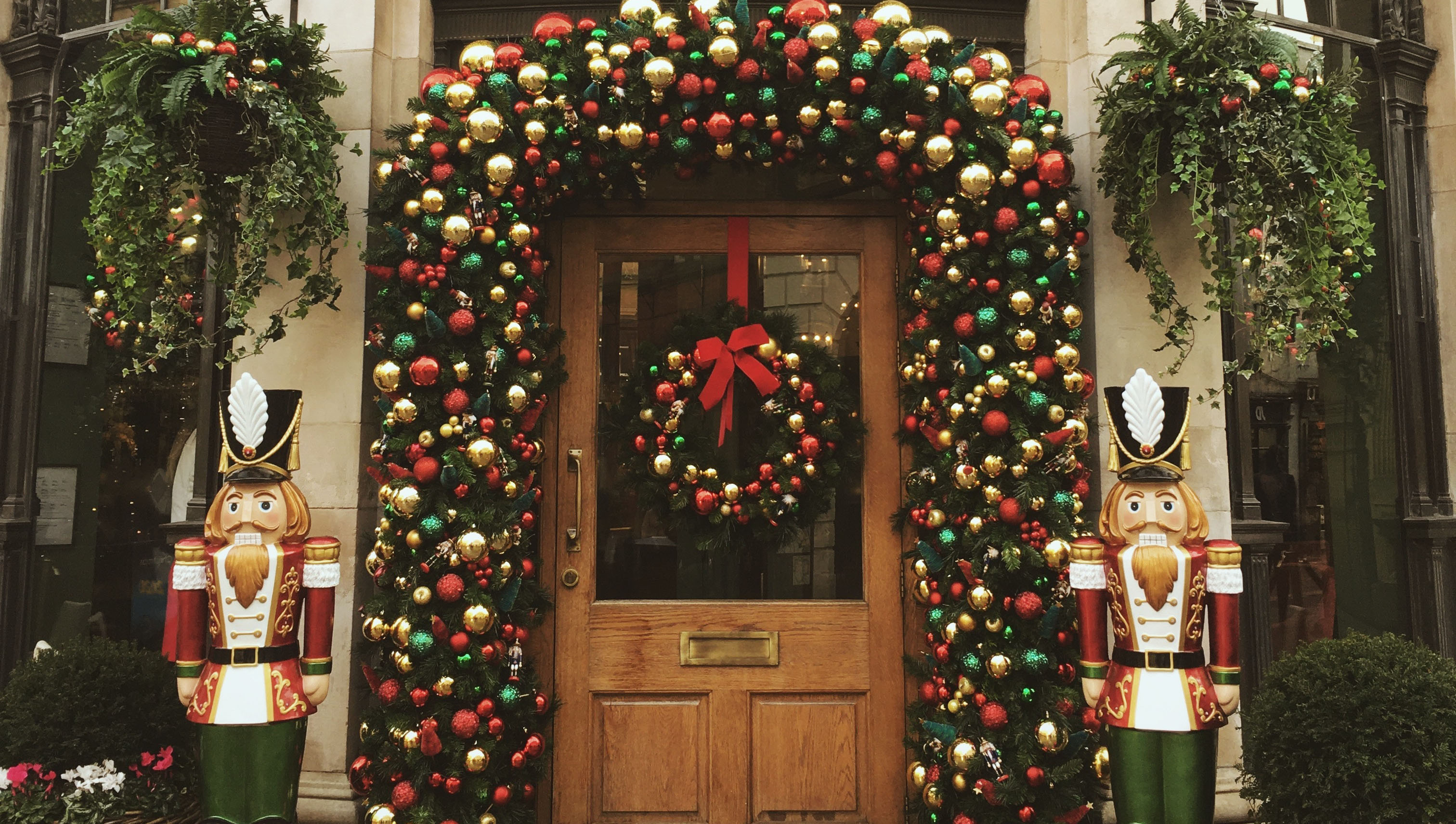 A festive doorway adorned with a lush arch of red, green, and gold ornaments. A wreath with a red bow hangs on the wooden door. Two large nutcracker statues stand on either side. Ferns in hanging baskets flank the doorway, enhancing the holiday ambiance.