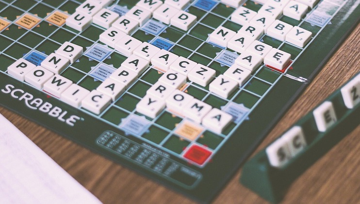 A Scrabble game board displays various words in a different language, with tiles forming words across and down. The word SCRABBLE is printed on the side of the board. A rack of tiles and a part of a score sheet are also visible.