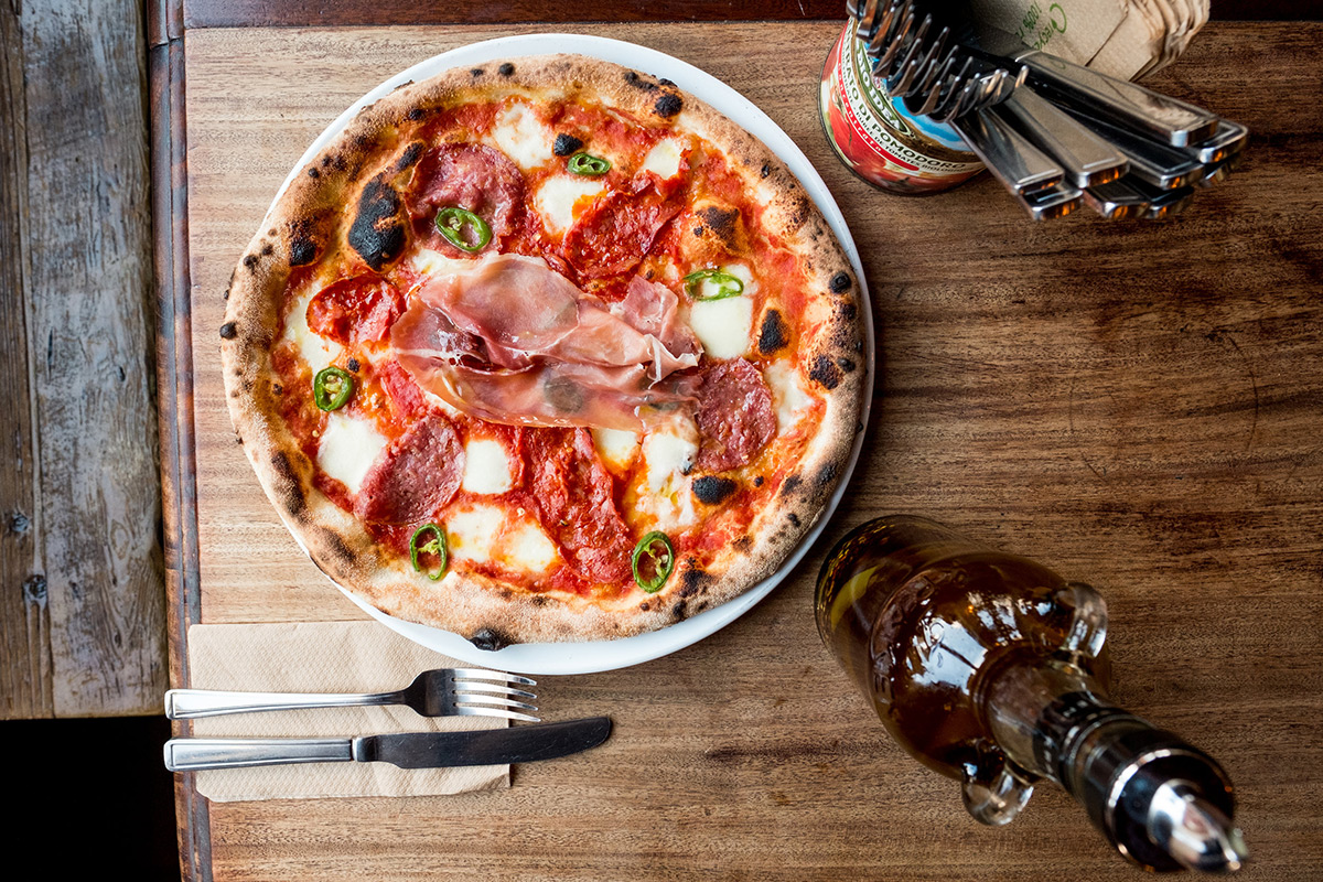 A wood-fired pizza topped with mozzarella, salami, prosciutto, and green peppers sits on a wooden table. Beside it are a knife and fork on a napkin, a bottle of olive oil, and a container with utensils.