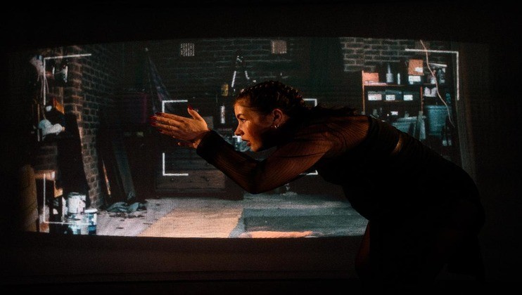 A person stands indoors, slightly bent over and clapping their hands together. They are dressed in dark clothing. Behind them, a screen displays a dimly lit room with brick walls, various shelves, and scattered items, creating a contrast between the real and projected spaces.