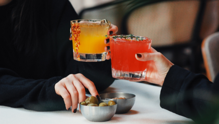 Two individuals holding colorful cocktails; one drink is yellow with a pepper garnish, while the other is red with a sprig of thyme. One person's hand is reaching for olives in a small bowl. Both are in a relaxed setting, with a white tablecloth underneath.