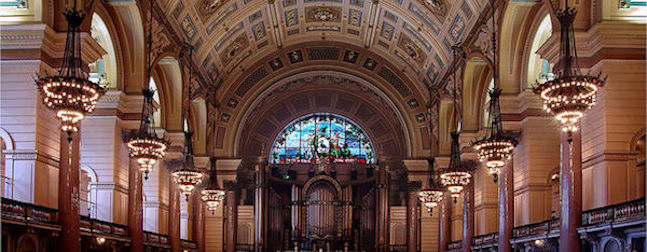 A grand, ornate hall with arched ceilings, stained glass windows, and large chandeliers. The space features a decorative organ and elaborate architectural details, creating an opulent and historic atmosphere.