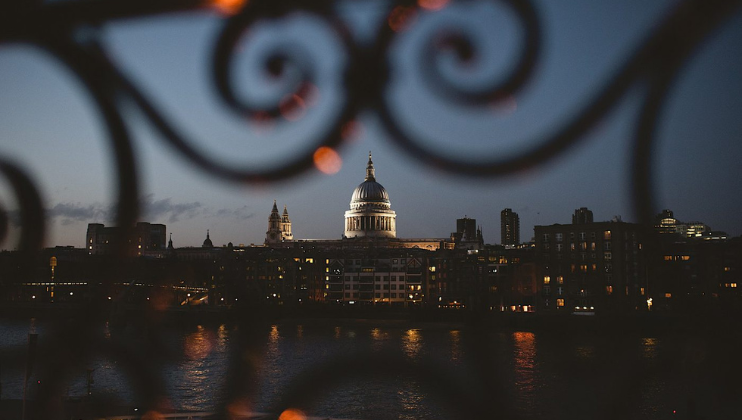 A view of a city skyline at dusk, with the illuminated dome of a prominent building centered in the background. The scene is framed artfully through blurred, decorative ironwork in the foreground, adding a touch of elegance to the image.