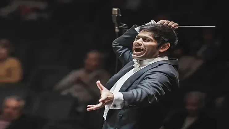 A conductor passionately leads an orchestra, his right arm raised with a baton in hand, and his left arm extended outward. He wears a dark suit with a white dress shirt. The background is blurred, featuring an audience seated in a dimly lit venue.