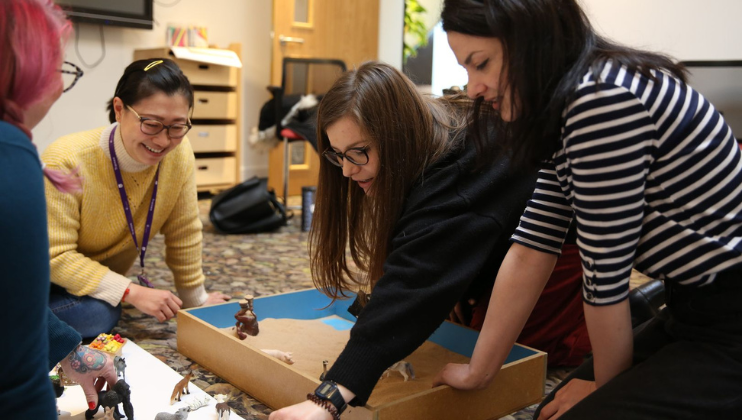 Four people are engaged in a table game, gathered around a rectangular board with game pieces. They appear to be focused and joyful. The setting looks like a casual indoor space, with bookshelves and a carpeted floor.