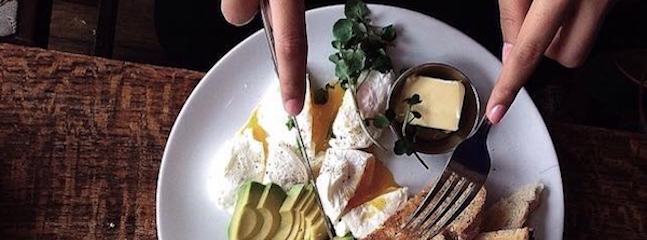 A white plate with a breakfast meal, including poached eggs, sliced avocado, butter, and toast. Two hands are visible, one holding a fork and the other pointing at the food. The plate is on a wooden table.
