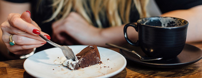 A person with red nail polish uses a fork to eat a slice of chocolate cake with cream on a white plate. A black coffee cup sits on a saucer beside the plate on a wooden table. The background is slightly blurred.
