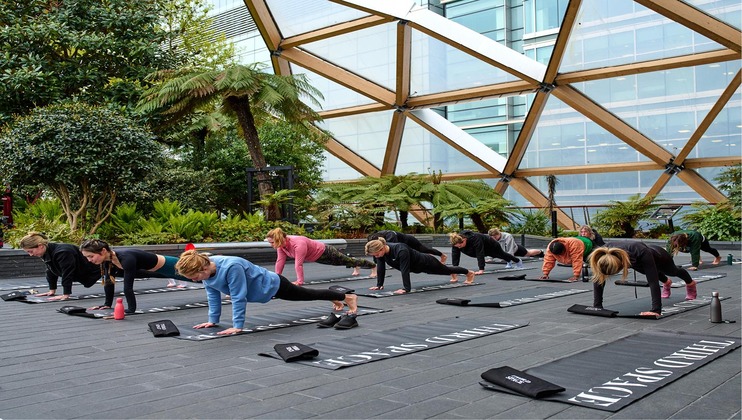 A group of people is practicing yoga on black mats in an outdoor setting surrounded by greenery and modern architecture. They are in a plank position, aligned in rows under a large glass structure with wooden beams.