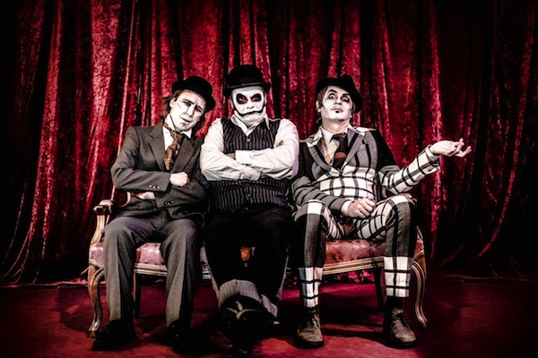 Three people in theatrical makeup and costumes sit on a vintage sofa against a red velvet curtain background. The person on the left wears a suit and bowler hat, the middle person wears a pinstripe suit and bowler hat, and the person on the right has a plaid suit.