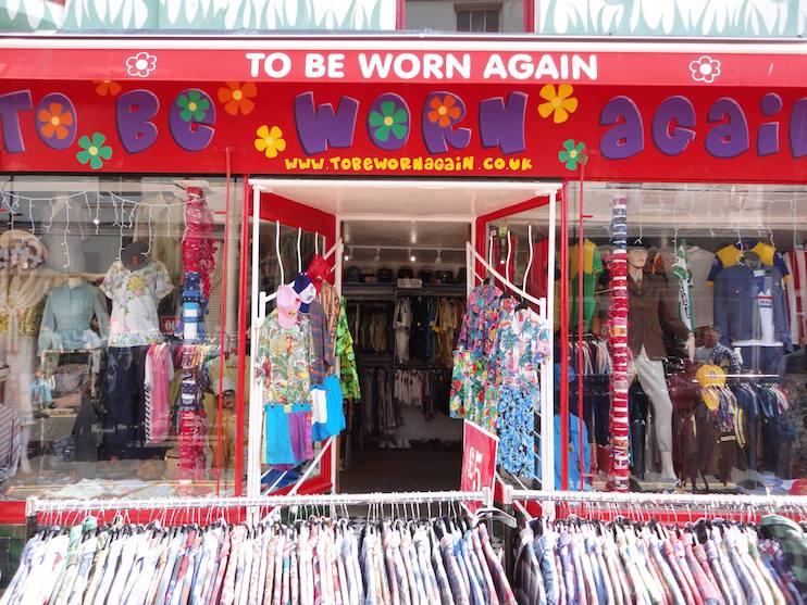 A vintage clothing store with a bright red sign that reads TO BE WORN AGAIN. The store window displays colorful clothing, and the entrance is open, revealing more vintage clothes inside. A website URL is displayed under the store name.