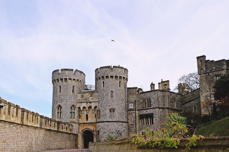 A historic stone castle with two round towers and an arched entrance stands under a partly cloudy sky. The walls are fortified and a small airplane is visible in the sky above. Some greenery and plants can be seen in the foreground.
