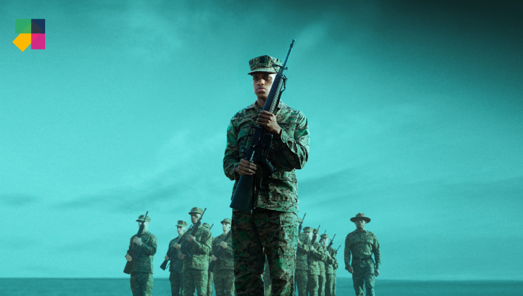 A soldier in camouflage uniform stands holding a rifle against his chest, with a group of similarly dressed soldiers standing in formation behind him. The background features a clear sky and some visible portions of landscape. Colored geometric shapes are seen in the top left corner.