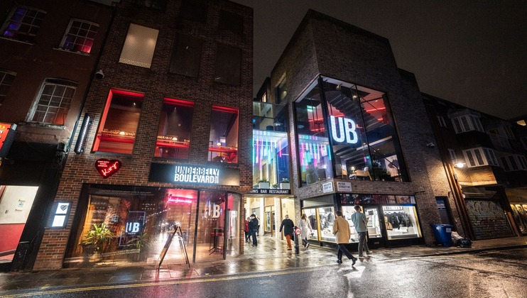 A nighttime street scene showing a modern building with large glass windows, neon signs, and people walking. The building is illuminated and features the sign URBAN BOULEVARD with glowing letters UB on the upper floors. The wet pavement reflects the lights.
.