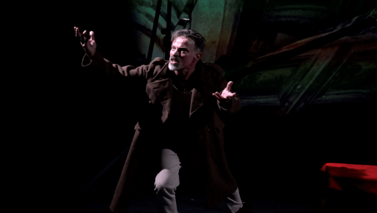 A person with gray hair and a beard is passionately acting on a dimly lit stage. They are wearing a brown coat and raising their arms dramatically. The background is dark with some indistinct shapes and colors.