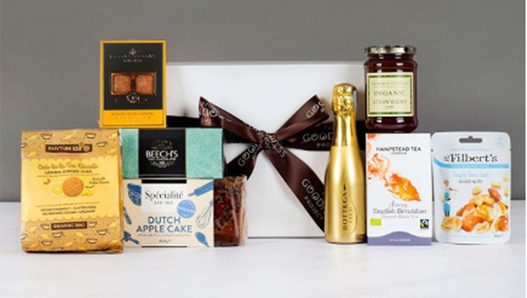 A selection of gourmet food items is displayed in front of a white gift box tied with a brown ribbon. The assortment includes cookies, chocolates, cake, jam, a small bottle of sparkling wine, tea, and nuts. The items are neatly arranged on a gray and white backdrop.