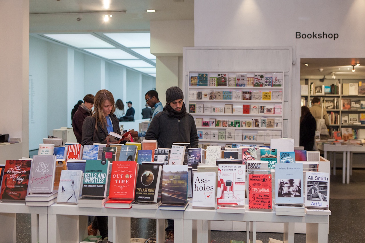 A modern bookshop features several shelves filled with various books. Two people stand near the front display, browsing different titles. In the background, other customers explore more books. The setting is well-lit with a clean and organized layout.