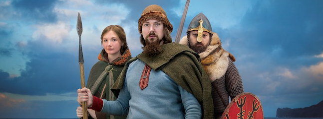 Three individuals in Viking attire pose against a backdrop of a cloudy sky and seascape. The central figure holds a spear and wears a fur-lined hat, flanked by a woman holding a spear on the left and a helmeted man with a shield on the right.