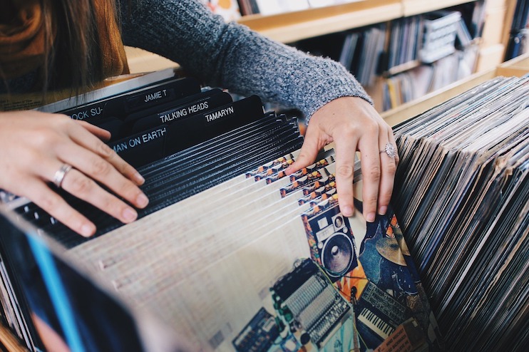 A person in a gray sweater flips through vinyl records in a wooden crate at a record store. The covers of records like Talking Heads and various others can be seen. The person is wearing several rings on their fingers.