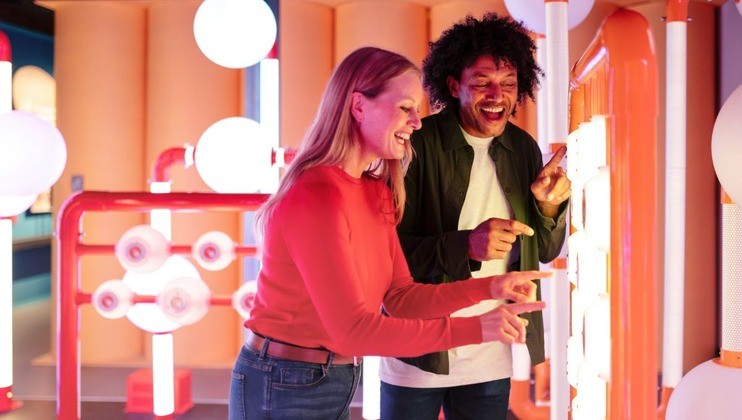 Two people enjoying an interactive exhibit in a brightly lit room with colorful lights and pipes. One person is wearing a red sweater, and the other is in a white shirt and dark jacket. Both are smiling and engaged with the exhibit.