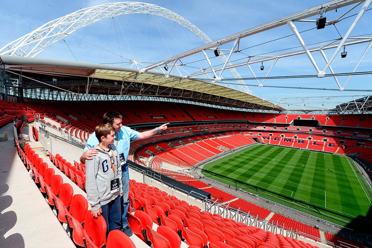 Two people, one adult and one child, are standing in the stands of a large, empty stadium. The adult is pointing towards the field, and both are looking in that direction. The stadium seats are red, and the field is green and well-maintained.