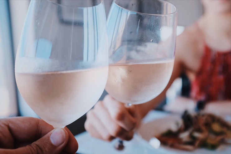 Two people toast with glasses of chilled white wine. One person is partially visible in the background, wearing a red top, and there is a meal on the table in front of them. The scene is softly lit, creating an intimate atmosphere.