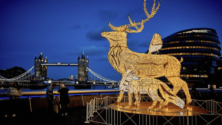 A night view of illuminated deer sculptures on display by the Thames River in London. The backdrop features the iconic Tower Bridge and modern glass buildings, with a few people admiring the artwork. The sky is a deep blue, adding a serene ambiance to the scene.
