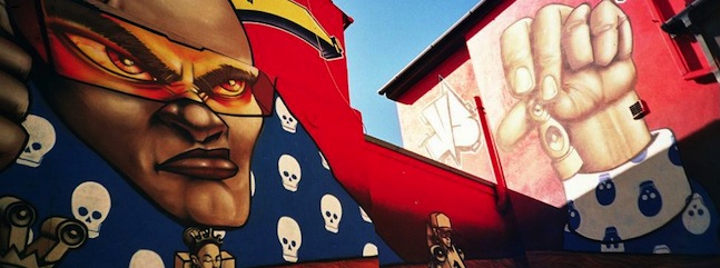 A vibrant street mural featuring a superhero with an intense expression, wearing a red mask and suit. The surrounding wall displays skulls and a close-up of a clenched fist holding a small object. Bright red and blue colors dominate the artwork.